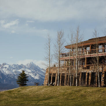 Amangani residence set on a hillside with snow-capped mountains in the distance, tall bare trees and alpine meadow in foreground.
