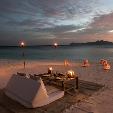 Private dining setup on Amanpulo's beachfront at dusk, with lanterns illuminating a wooden table on the sand.