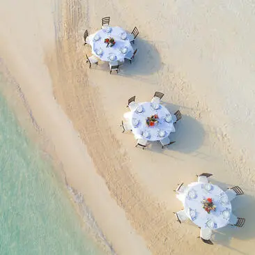 Amanpulo resort এ সমুদ্র সৈকতে সন্ধ্যাকালীন বিবাহ অনুষ্ঠানের জন্য সাদা ছাতা এবং টেবিল সহ তিনটি ডাইনিং সেটআপ।