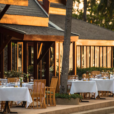Tavoli all'aperto del ristorante Thai di Amanpuri al tramonto, con luci calde e vegetazione sullo sfondo.