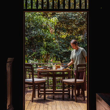 Woman seated at wooden dining table on terrace at Amansara, framed by doorway with garden view beyond.