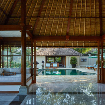 Open-plan living area at Amandari with thatched roof, wooden beams and view to pool beyond.