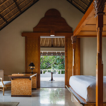 Amankila suite interior with wooden furnishings, peaked ceiling, and open terrace doors overlooking tropical gardens.