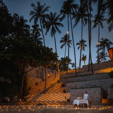 アマンプリ・リゾートの夕暮れ時の庭園。石造りの壁とヤシの木に囲まれた照明されたテラス。
