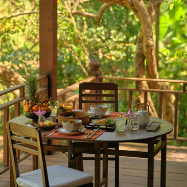 Breakfast table set on a wooden deck at Amansara, surrounded by lush greenery and natural light filtering through trees.