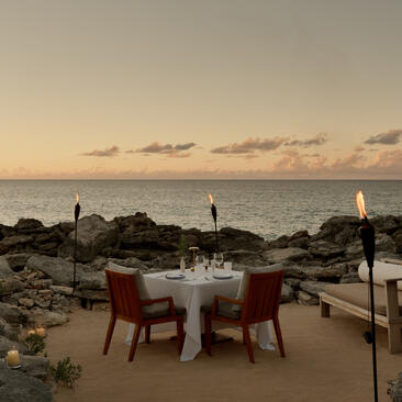 ターコスカイコス諸島のアマンヤラで、ビーチコーブでの二人席の食事体験。夕暮れ時、白いテーブルと赤いチェアが岩場に置かれ、静寂の海を望む。