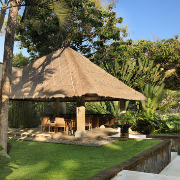 Thatched-roof dining pavilion at Aman Villas at Nusa Dua, surrounded by tropical gardens and palm trees.