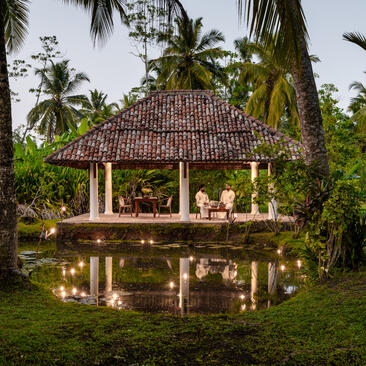 Amangalla's traditional Sri Lankan pavilion reflected in a still pond, surrounded by lush tropical gardens and palm trees.