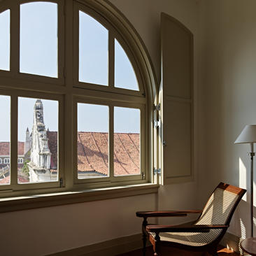 Arched window framing colonial architecture and terracotta rooftops at Amangalla, with a wooden chair beside.