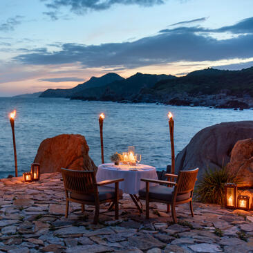 Two chairs and a fire pit on a beach at Amanoi at dusk, with mountains across the water.