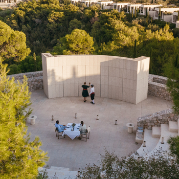 Aerial view of a stone amphitheatre dining venue at Amanzoe resort, surrounded by Mediterranean landscape and pine trees.