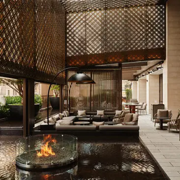 Terraço ajardinado do hotel Aman New York com lareira, elementos de madeira e vidro, refletindo-se na água.