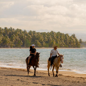 Two riders on horseback walking along a sandy beach with forested coastline at Amanera, Dominican Republic.