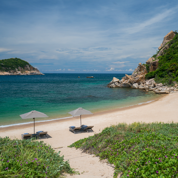 Wooden pathway leading through coastal vegetation to a sandy beach at Amanoi, with turquoise waters and moored boats.