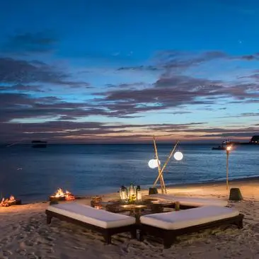 Cena privata sulla spiaggia al tramonto presso il resort Amanpulo, con tavoli bassi, lanterne e fuoco sulle sabbie tranquille.