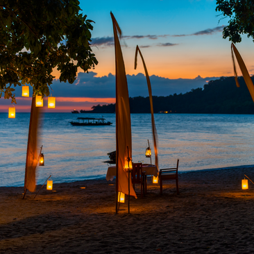 Candlelit private dining setup on Amanwana's beach at sunset, with illuminated sail structures and ocean view.