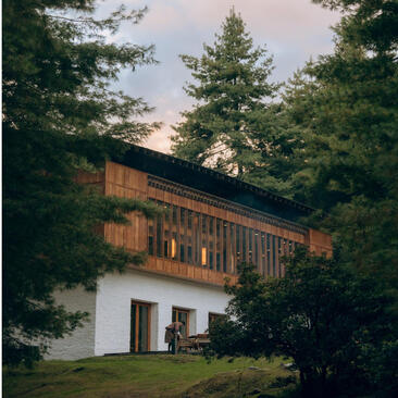 Amankora lodge in Paro nestled among pine trees at dusk, with traditional Bhutanese architecture featuring wooden upper floors and whitewashed base.