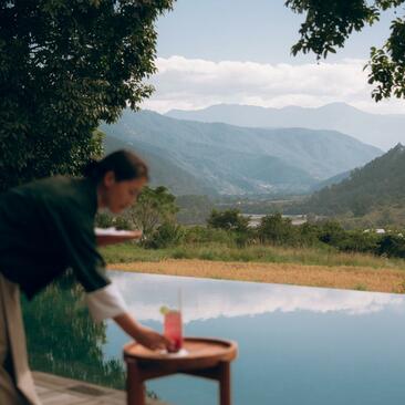 Man reading at a wooden table overlooking a plunge pool, with Punakha valley mountains beyond at Amankora.