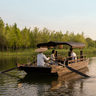 Traditional wooden boat on West Lake at Amanyangyun, with guests seated beneath a canopy whilst gliding past green shoreline.