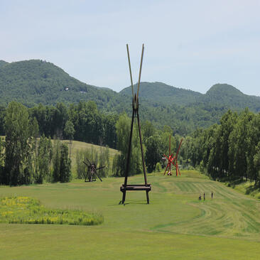 Tall sculptural installation on manicured lawn at Aman New York, with tree-lined grounds and mountains beyond.
