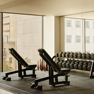 Bright fitness studio at Aman New York with exercise equipment and a quilted seating area near windows overlooking the city.