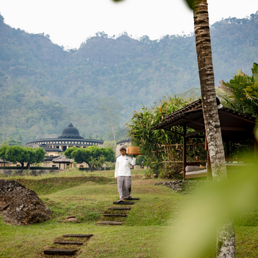 A figure walks across manicured grounds towards a temple at Amanjiwo, with forested mountains rising beyond.