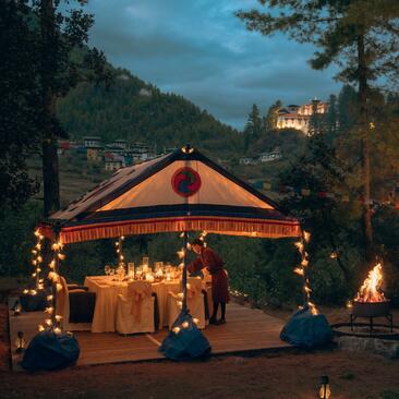Candlelit dinner pavilion in Paro forest at dusk, with illuminated fortress visible on hillside above.