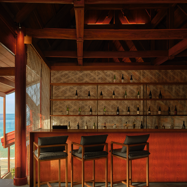 Nama sake bar setup at Amanpuri resort in Thailand, featuring wooden beams, terracotta walls, and shelved spirits display behind bar seating.