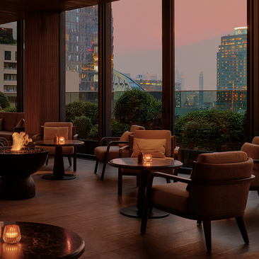 Aman Nai Lert Bangkok lounge at dusk, with floor-to-ceiling windows overlooking Bangkok's skyline and candlelit seating areas.