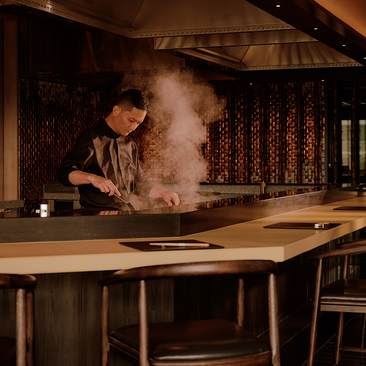 Chef preparing a dish with theatrical flame at Aman Nai Lert Bangkok's dining venue.