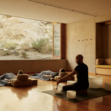 A person sits cross-legged on a wooden floor during a sound bowl wellness experience at Amangiri, with a large landscape artwork on the wall.