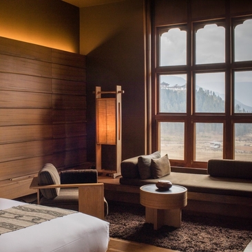 Gangtey Lodge suite bedroom at Amankora with wooden walls, seating area by arched windows overlooking the valley, and warm lighting.