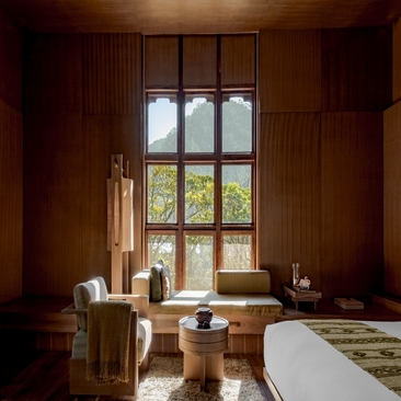 Warm-toned principal bedroom at Amankora's Punakha Lodge with wooden panelling, tall windows overlooking valley views, and freestanding bathtub.