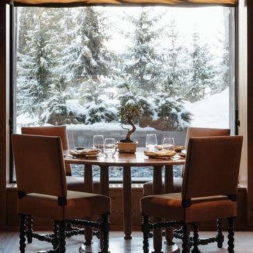 Dining table set for two beside a window overlooking snow-covered pine trees at Aman Le Mélézin.