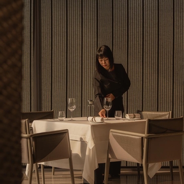 曼谷奈乐安缦酒店内，一位女性在餐桌前用餐，温暖灯光映照米色织物和竖条纹背景。