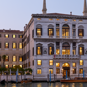 Aman Venice's palazzo illuminated at night, its yellow façade reflected in the Grand Canal waters below.