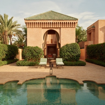 Moroccan pavilion with terracotta walls reflected in still pool water at Amanjena, surrounded by manicured hedges and palm trees.