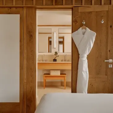 Petite chambre avec murs en bois clair, lit blanc et robe de chambre suspendue, Aman Le Mélézin.