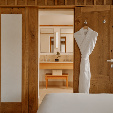 Small bedroom at Aman Le Mélézin with wooden walls, simple furnishings and a white robe hanging on the door.