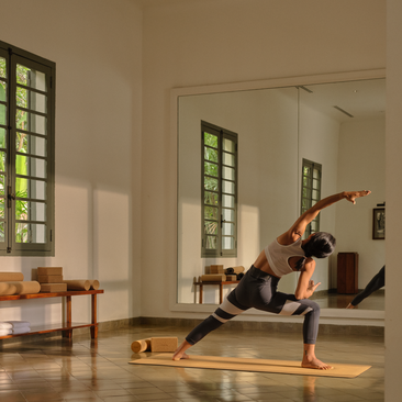 Yoga instructor leading a sunrise movement class at Amantaka, demonstrating a standing pose on a wooden platform in a light-filled studio.