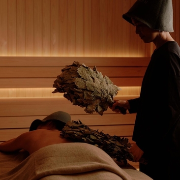 Therapist performing herbal bundle treatment on guest in wooden sauna at Aman Nai Lert Bangkok wellness spa.