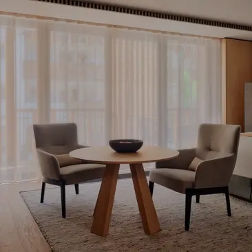 Deluxe Room with two grey chairs facing a round wooden table at Aman Rosa Alpina resort, sheer curtains filtering natural light.