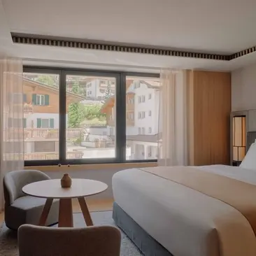 Superior Room 211 at Aman Rosa Alpina resort, featuring a bed, round table by windows overlooking the Italian Alps, and wooden wardrobe.