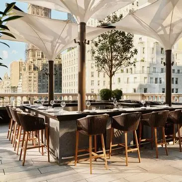 Terraço ao ar livre do bar no Aman New York com mesas e cadeiras de madeira, guarda-sóis brancos e vista para a cidade.