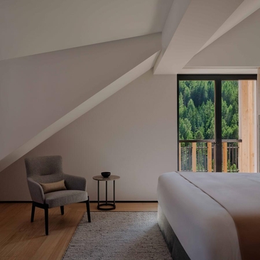 Dolomites Suite bedroom at Aman Rosa Alpina with angled ceiling, armchair, and mountain views through tall windows.