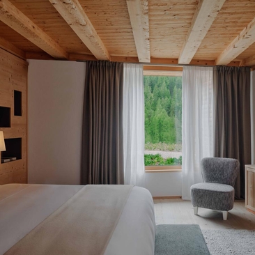 Zeno Suite principal bedroom at Aman Rosa Alpina with wooden ceiling, large windows overlooking Italian Alpine landscape, and contemporary furnishings.