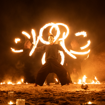 Silhouette of a figure twirling fire poi on Amanpuri's private beach at dusk, creating glowing arcs of light.