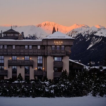 Aman Le Mélézin exterior at dusk, with snow-covered peaks glowing in alpenglow light beyond.