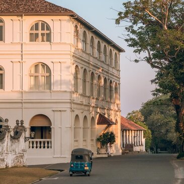 Exterior of Amangalla's colonial-era building with arched windows, a vintage tuk-tuk parked on the street, and Sri Lankan landscape beyond.