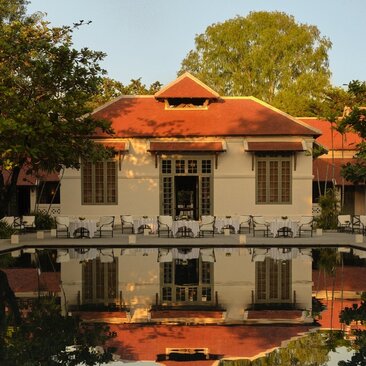 Amantaka's dining pavilion reflected in still water, with terracotta roof and colonial architecture framed by trees.